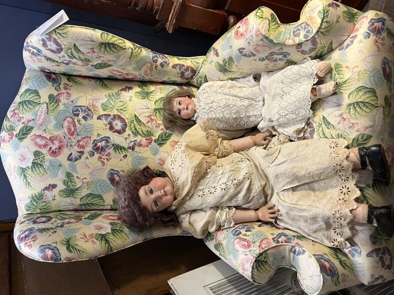 Two old dolls sitting on an antique chair, the dolls look quite creepy.