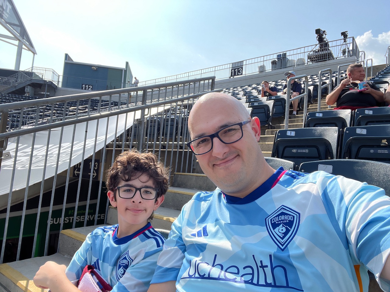 A kid and his parent, sitting in a stadium, wearing Colorado Rapids jerseys.